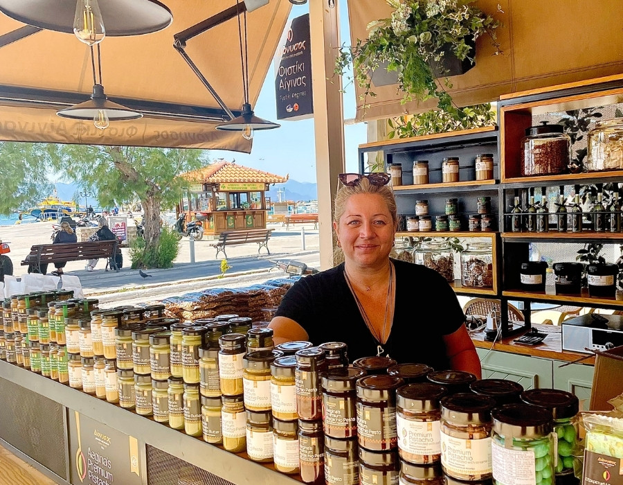 A woman stands in an open air shop with shelves of pistachio products, with the port beyond. The location is the Greek island of Aegina.