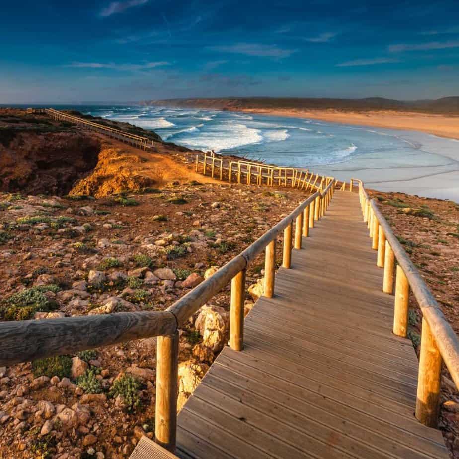 The Portugal coastline is pictured in the Alentejo region. A wooden boardwalk leads down towards the ocean. The blue sea with waves are pictured in the distance.