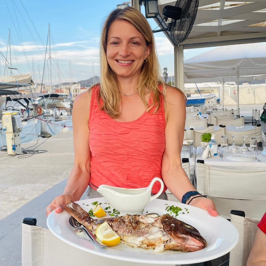 Ashley Blake, founder of Traverse Journeys holds a platter with a red snapper, on the Greek Island of Aegina, during a sailing trip