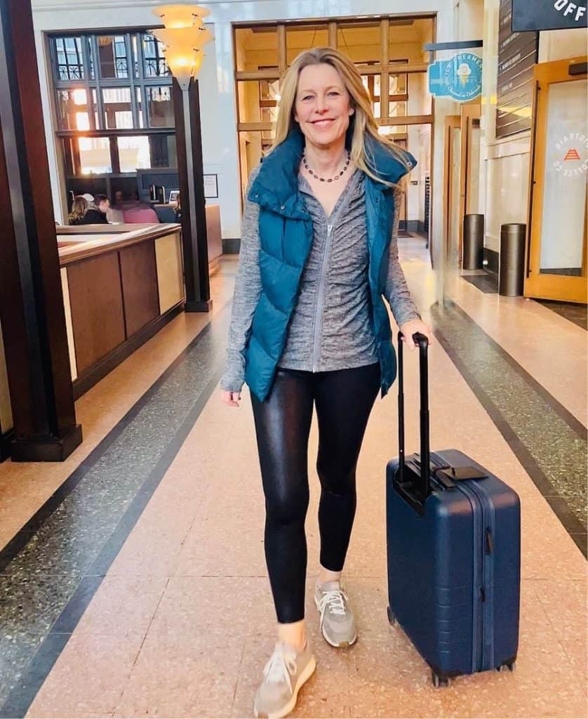 A woman is walking in a train station holding the handle of a suitcase, smiling. She has long blonde hair and is wearing black leggings, and a blue vest open at the front and a grey sweatshirt underneath. She is wearing sneakers.