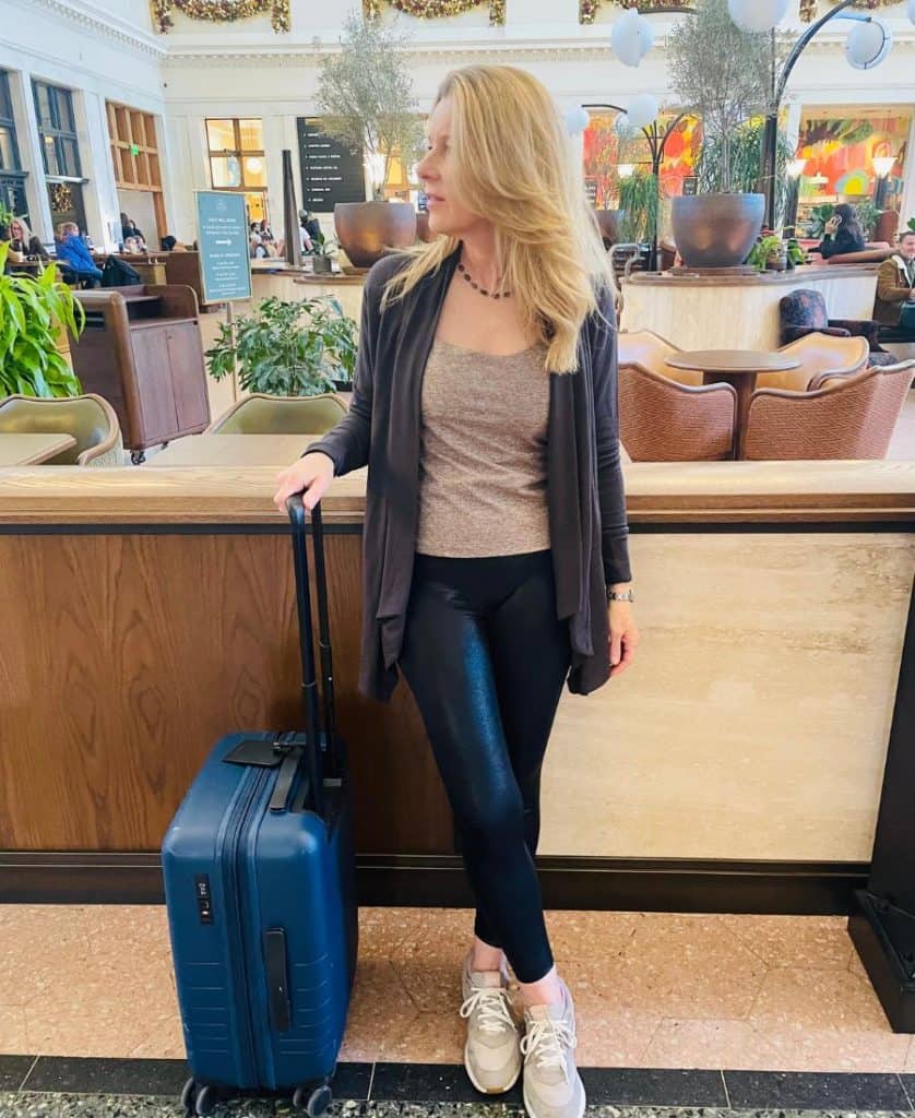 A woman is pictured in a train station holding the handle of a suitcase and looking off to the side, smiling. She has long blonde hair and is wearing black leggings, and a brown sweater open at the front. A light brown tank is visible underneath. She is wearing sneakers.