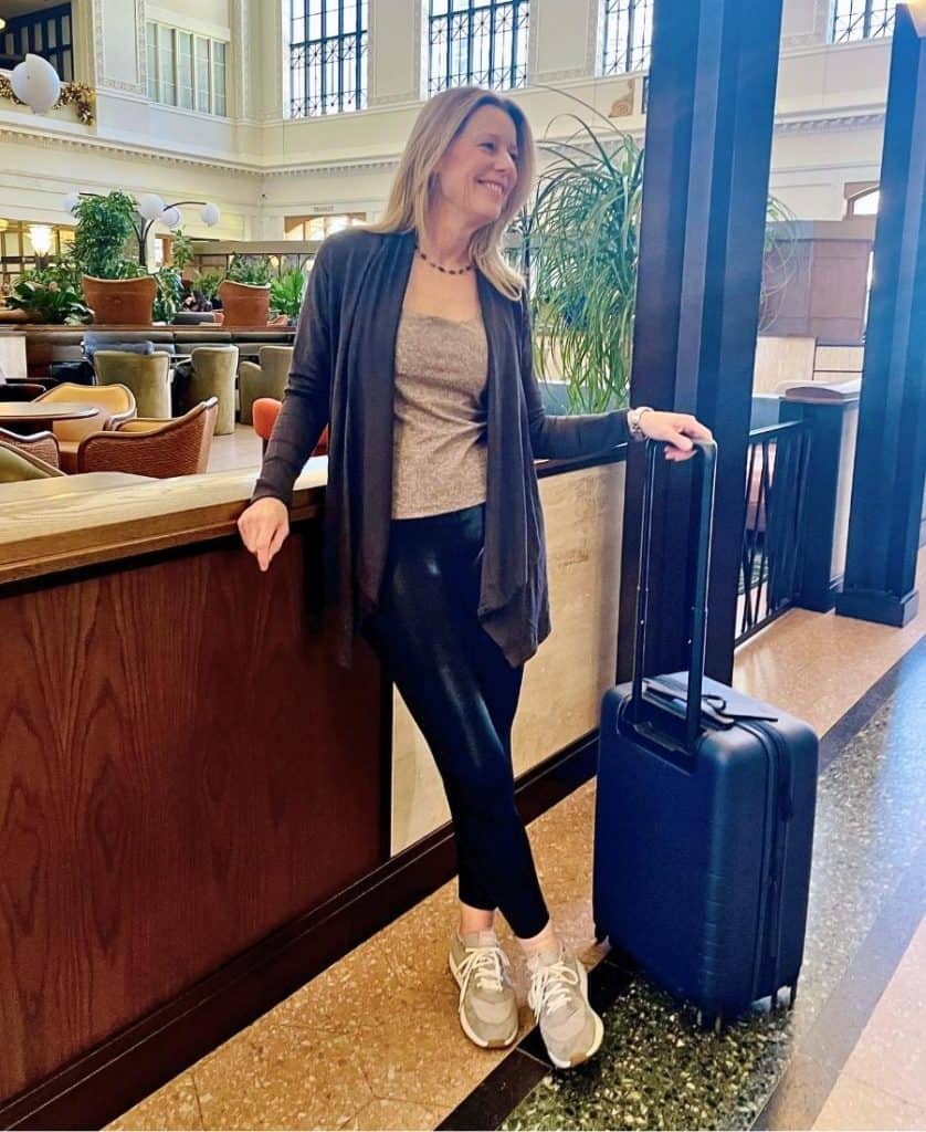 A woman is pictured in a train station holding the handle of a suitcase and looking off to the side, smiling. She has long blonde hair and is wearing black leggings, and a brown sweater open at the front. A light brown tank is visible underneath. She is wearing sneakers.