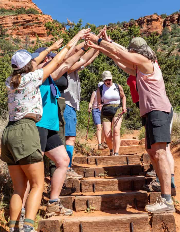 A group of women form an arch with their arms while other women walk through. They are in a red rock canyon.