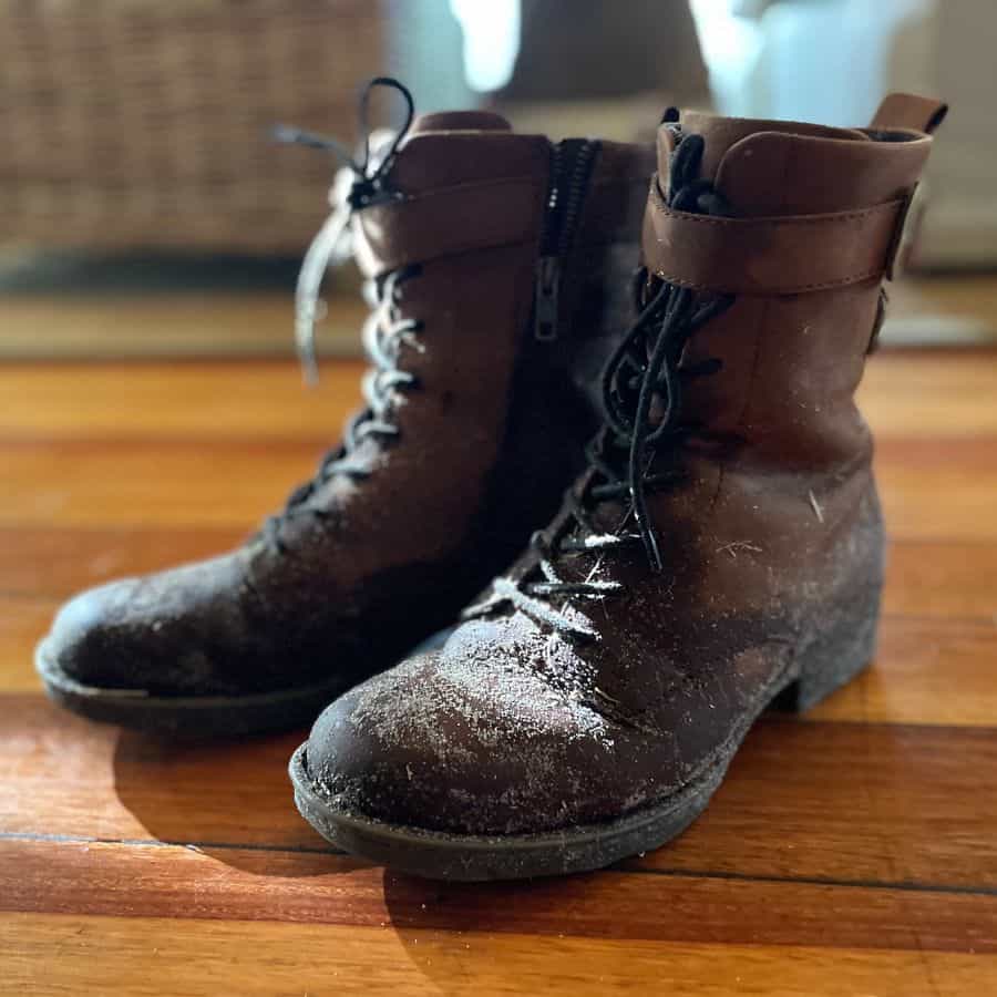 A pair of brown ankle boots are pictured, stained with mud and bits of grass. The are set on a wood floor with rattan furniture in the distance.