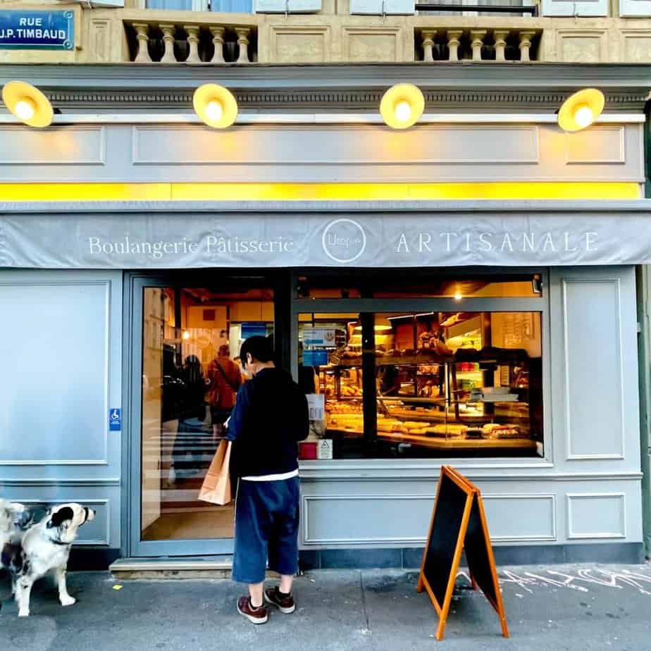 A pale-blue colored exterior of a bakery in Paris with an awning with gold-colored lights. A blackboard sign sits on the sidewalkand a man and a dog stand just outside the door. A warm light is emitted from inside.