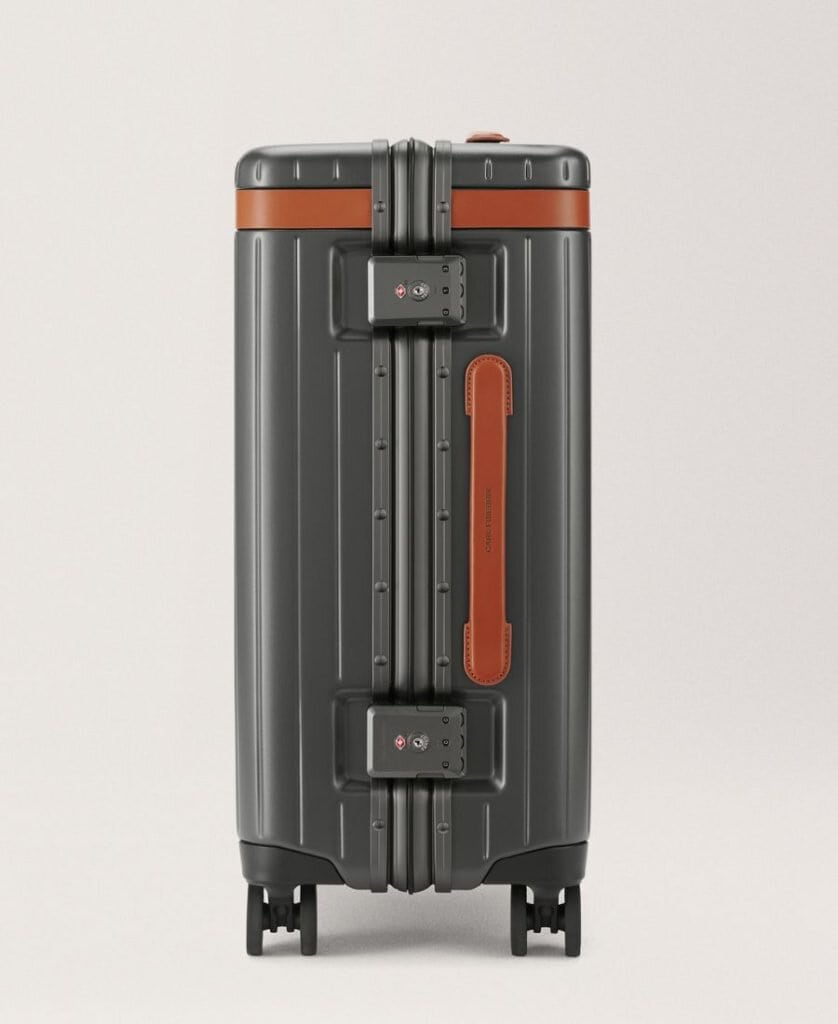 A carry-on roller bag is pictured from the side view. It is dark grey with brown leather trim and has four wheels. It has a plain background beyond it
