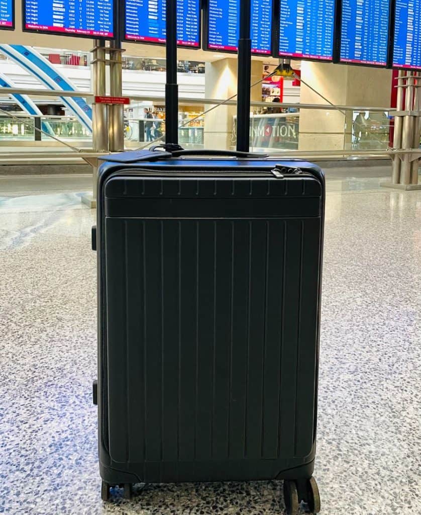 A small black roller bag stands with a digital flight board displayed beyond
