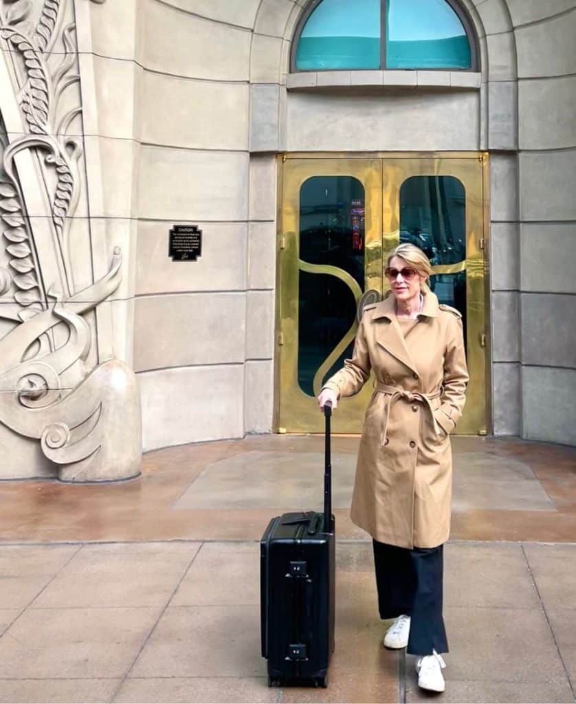 A blonde woman wearing black pants and a beige trench coat and sunglasses walks in front of what looks like a European building. She is holding the extended handle of a black carry-on roller bag.