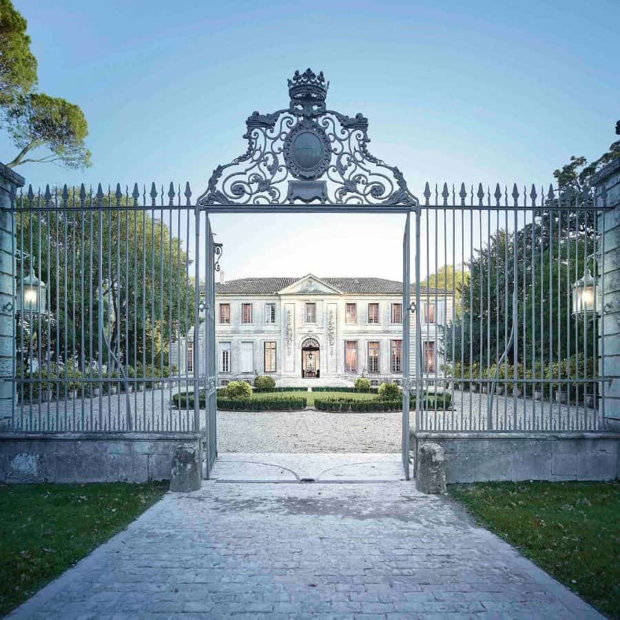 A historic French chateau is shown with an open gate and courtyard. Trees are on either side and a symmetrical garden at the center of the courtyard. Photo is by Brice Pelleschi.