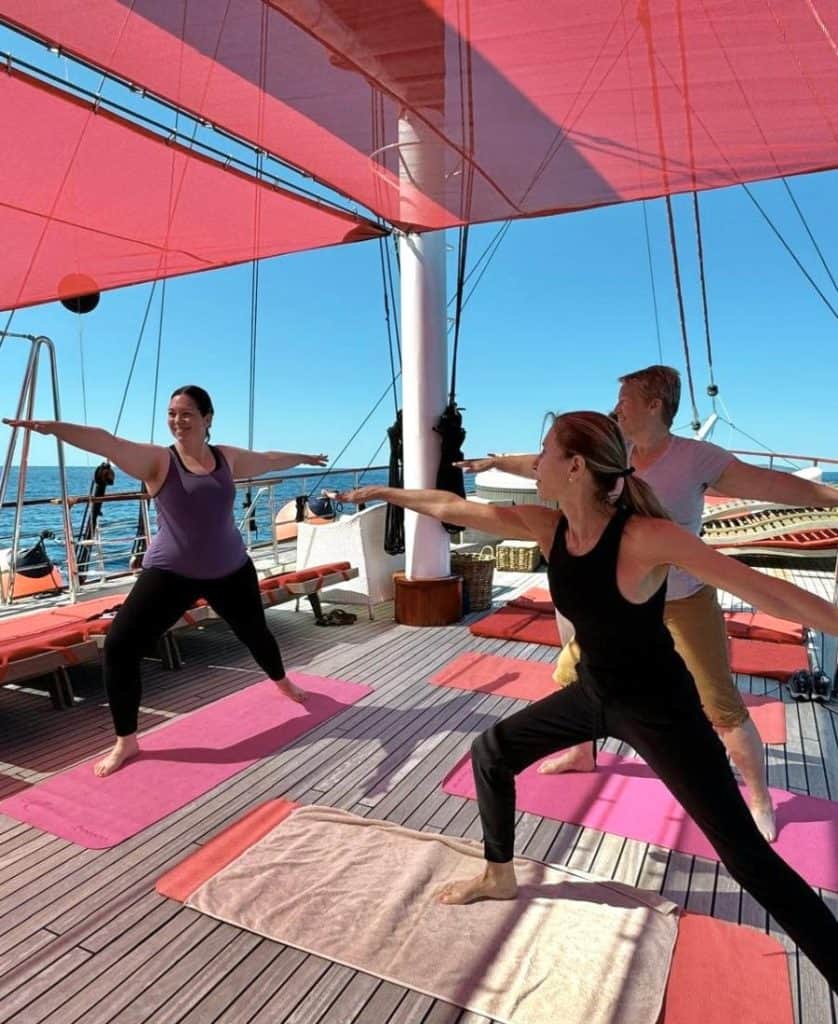 Three women are in yoga poses on a sailboat with red awnings over top of them.