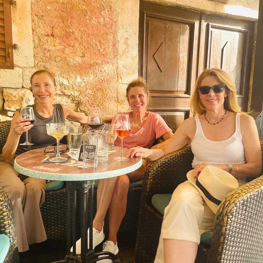 Three women sit at a small table with drinks on it. The background looks like an old building in Europe.