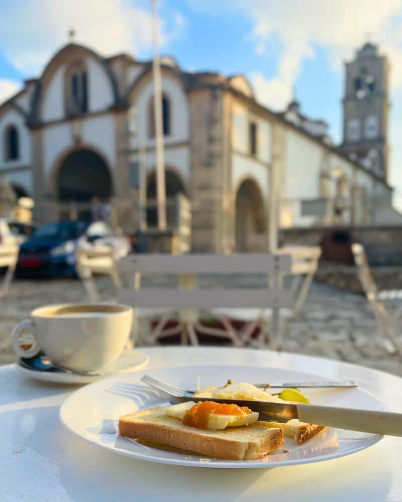A plate of breakfast and a cup of coffee are pictured atop a table on an open-air terrace. The facade of a 14th century church is visible across the square.