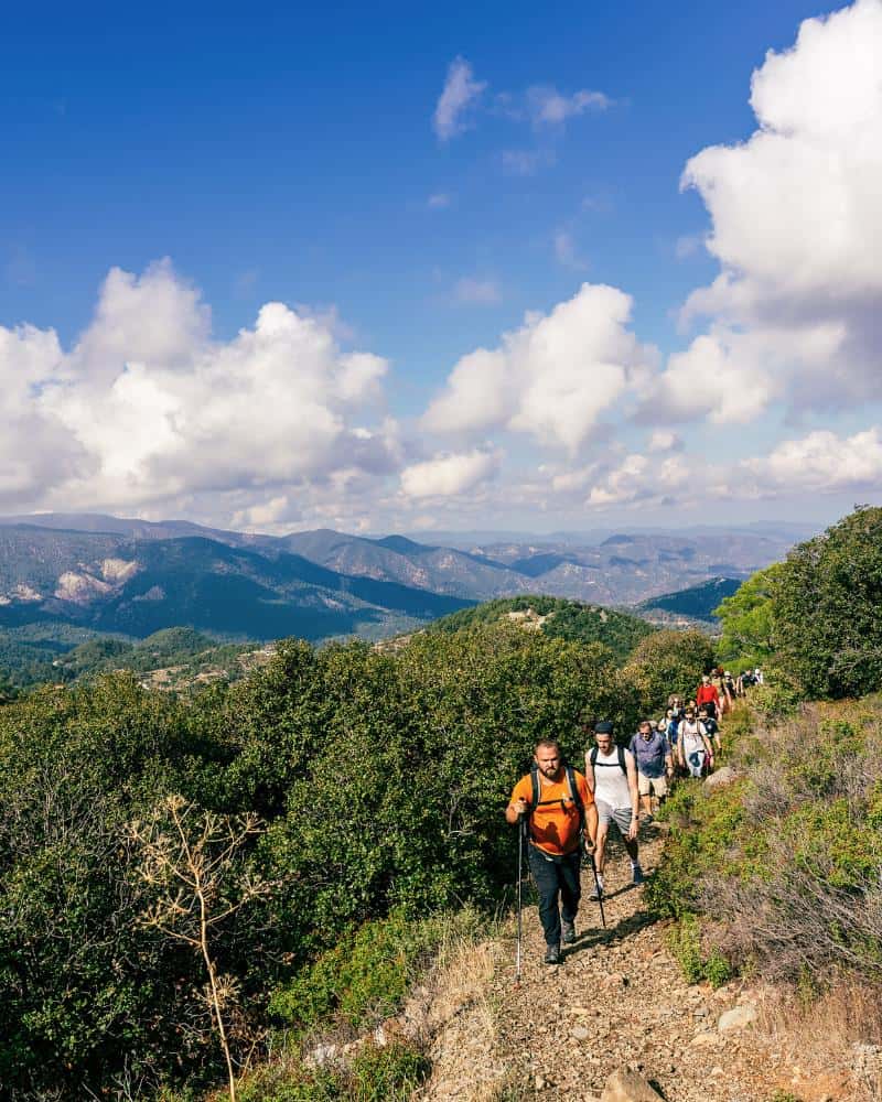 A line of people hike toward the camera along a narrow trail in the hills in Cyprus. The sky is blue with clouds beyond.