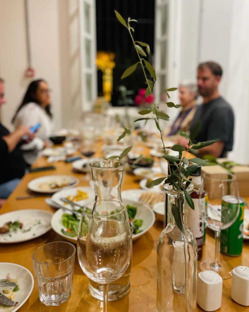 A wooden table is set with many dishes and diners are seen at the end of the table. Wine glasses and a small vase are in focus in the foreground.