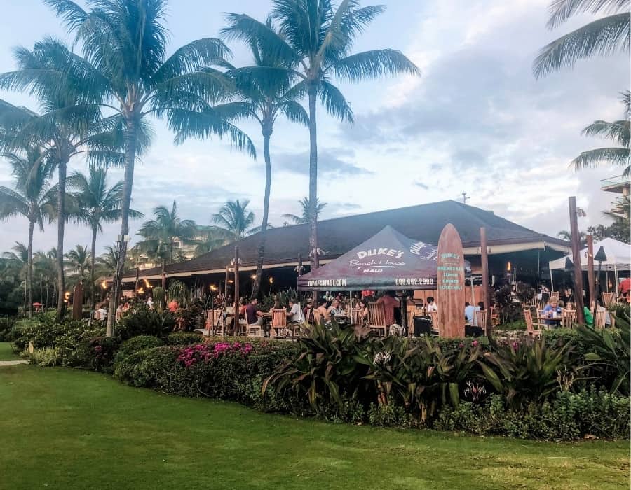 The exterior of Duke's restaurant on Kaanapali Beach in Maui. Tall palm trees are beyond the Polynesian-style restaurant lit with tiki torches.