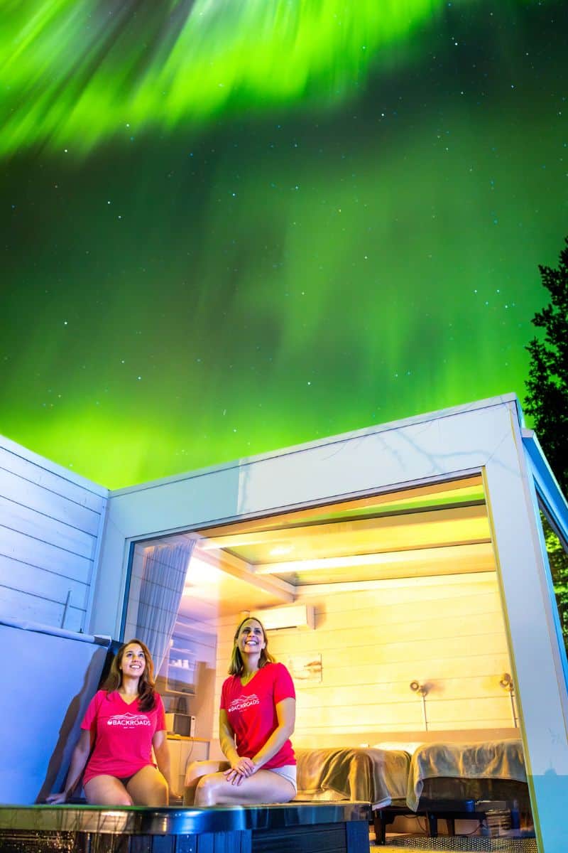 Two women sit in an outdoor hot tub with the northern lights in the sky above, in shades of green and gold.
