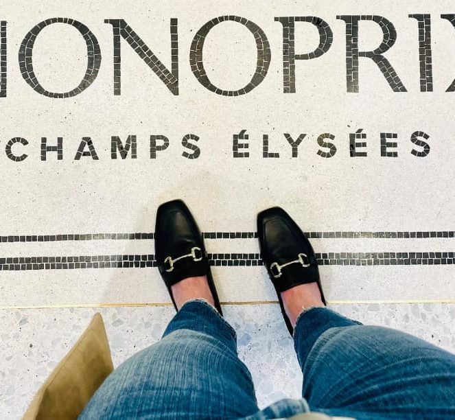 A woman's feet pictured in black French-style loafers on a tiled floor with the words: Monoprix Champs Élysées in decorative tile. Visible are her legs with blue denim and a beige trench coat. She is in Paris.