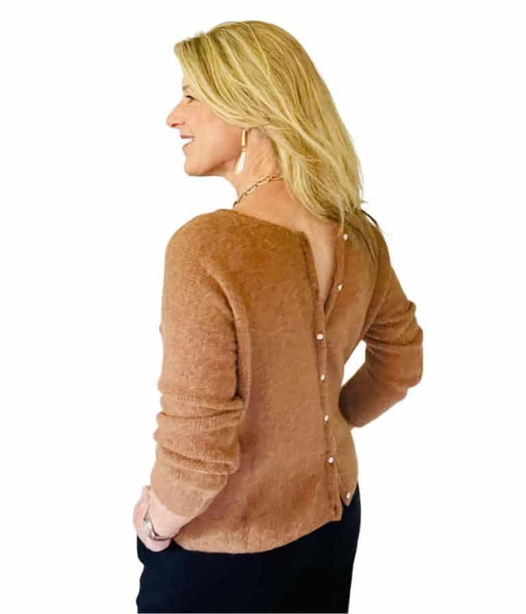 Susan Heinrich is wearing a camel-colored cardigan from Paris brand, Sezane. She is pictured from the side and also wearing black trousers. The cardigan is button at the back.