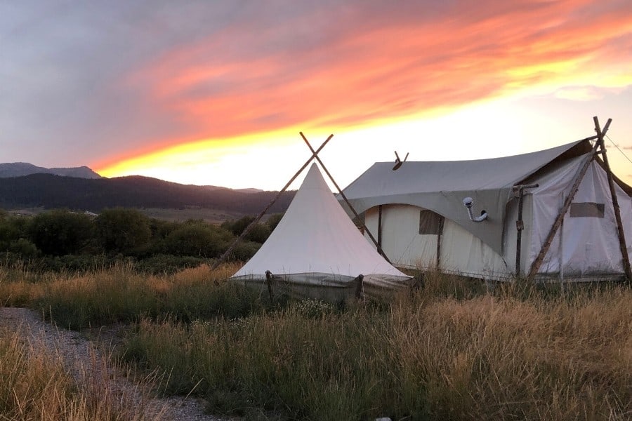 A white glamping tent and tipi are set in a mountain meadow with the sun setting beyond