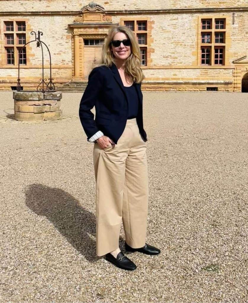 A woman stands in a courtyard in France wearing a classic French wardrobe: black Gucci-Style loafers, trousers and a navy blazer.