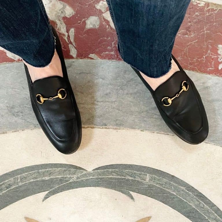 A pair of feet are pictured standing on patterned marble tile wearing traditional black loafers with a gold "horsebit"