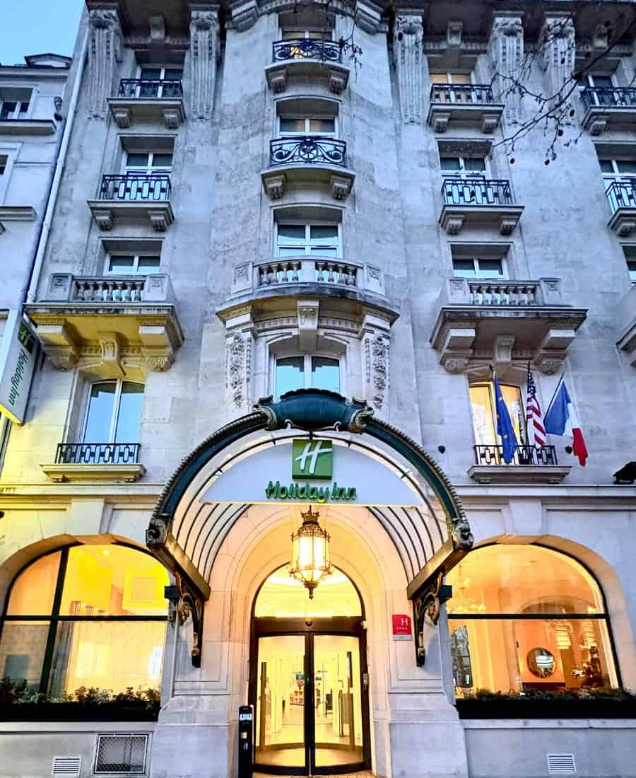 The exterior of a hotel in Paris in early evening, with traditional Haussmann architecture and a warmly lit entrance.