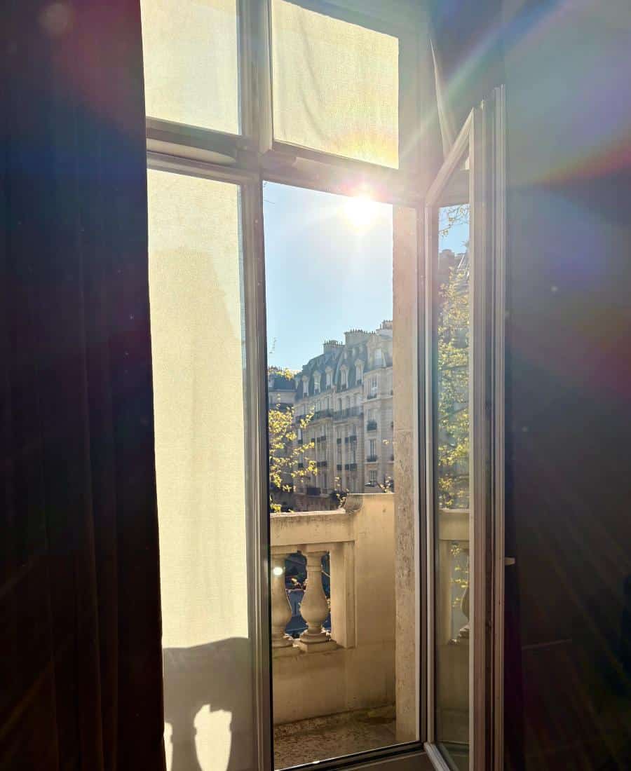 The view from a hotel room. Pictured is double French doors with the sun coming in. And a small balcony and leafy trees outside.