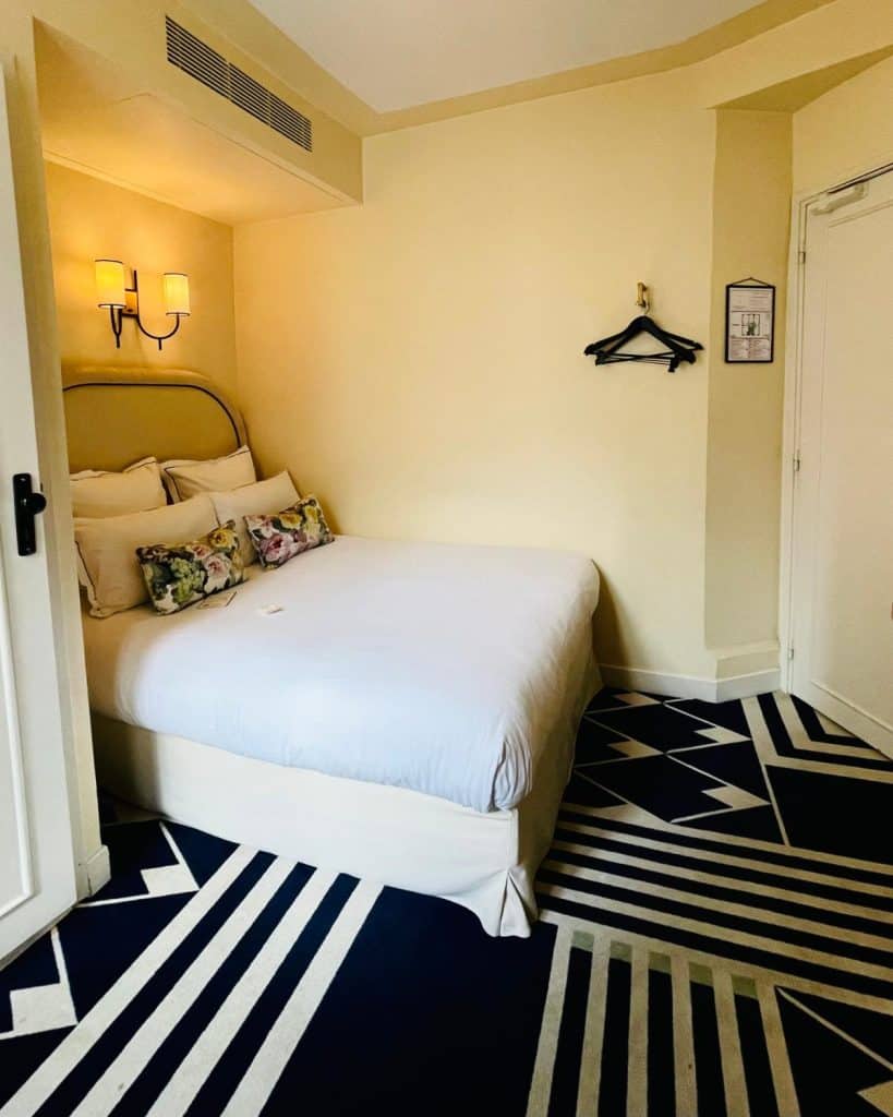 A stylish hotel room with a graphic black and white carpet and a double bed against the wall with white bedding and an upholstered headboard. A wall sconce light hangs above the bed.