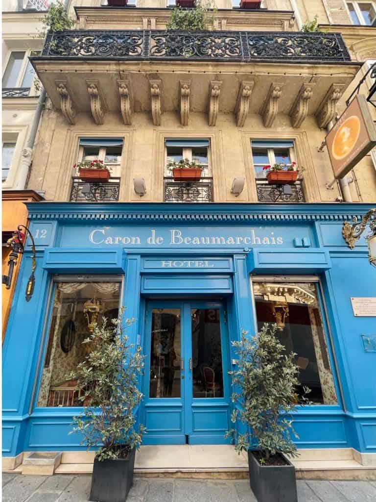 The exterior of the Hotel de Caron Beaumarchais in Paris. The first two floors are shown of a traditional Haussmann-style building, wit blue doors and flower boxes at the windows.