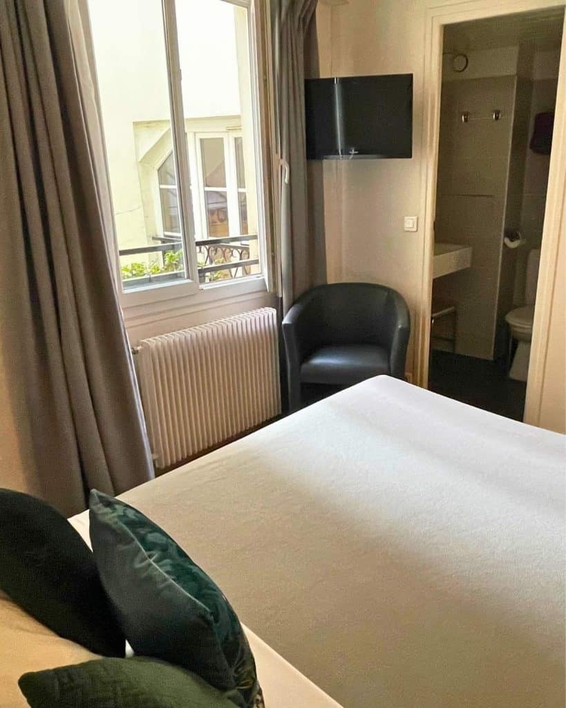 The interior of a Paris hotel room with simple decor, a double bed with green accent pillows, an armchair and a wall tv. An open window is visible as well as a door leading to a small bathroom.