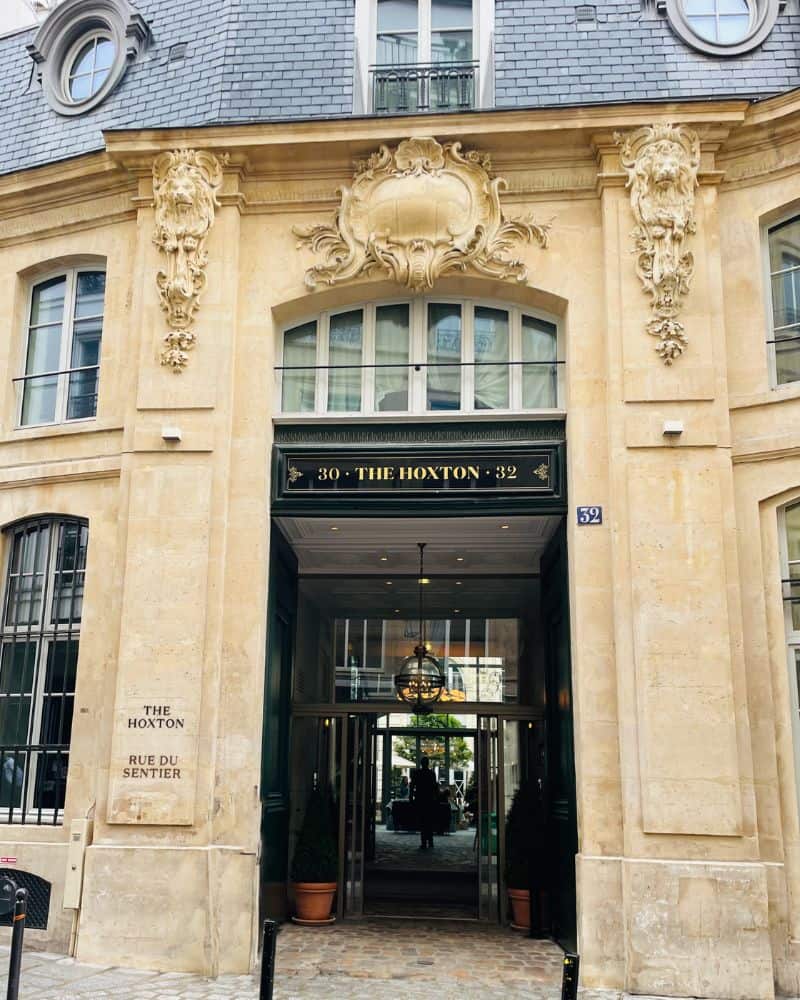 The exterior of a Haussmann-style building in Paris with an open entrance and door. Above the door are the words The Hoxton. There are cobblestones at the entrance. The building has some decorative features and a slate roof.