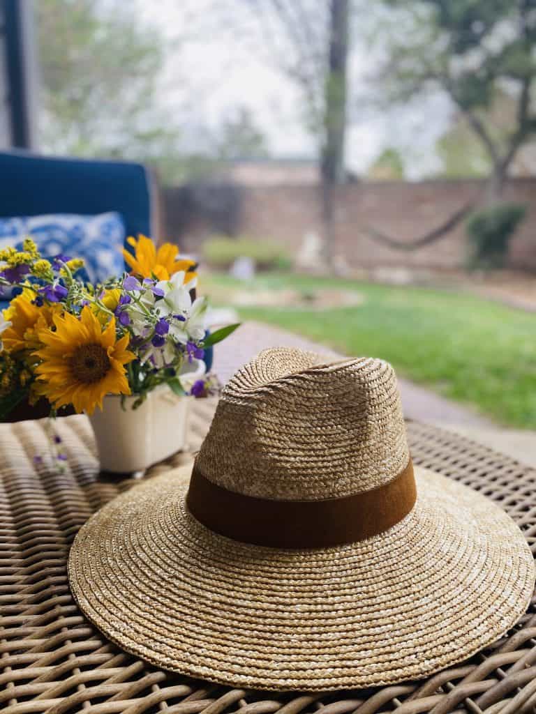 A straw fedora by Gigi Pip sits on an outdoor table next to a vase of wildflowers