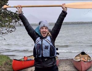 Kelly Kimple, CEO of Adventures in Good Company holds a paddle in the air. Behind her are two canoes on the beach of a lake.
