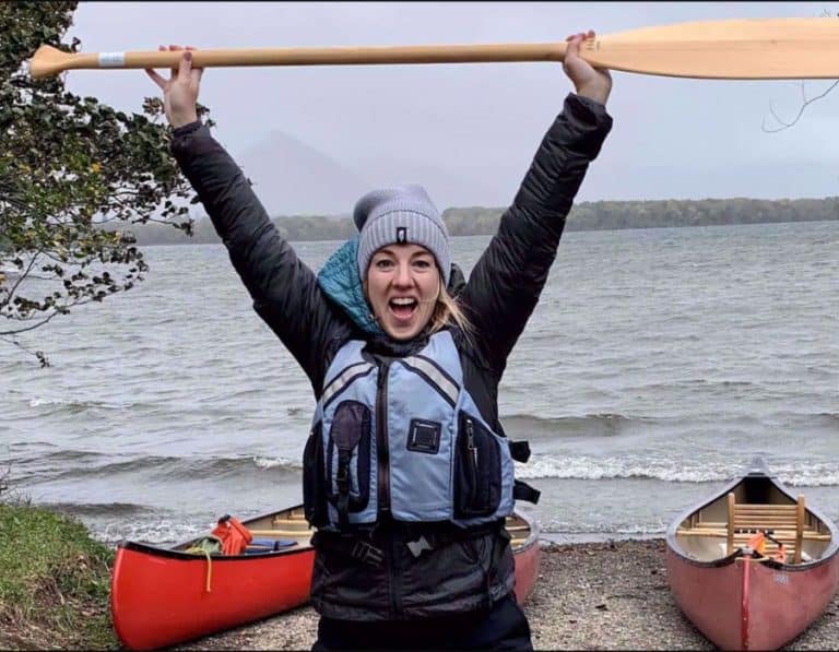 Kelly Kimple, CEO of Adventures in Good Company holds a paddle in the air. Behind her are two canoes on the beach of a lake.
