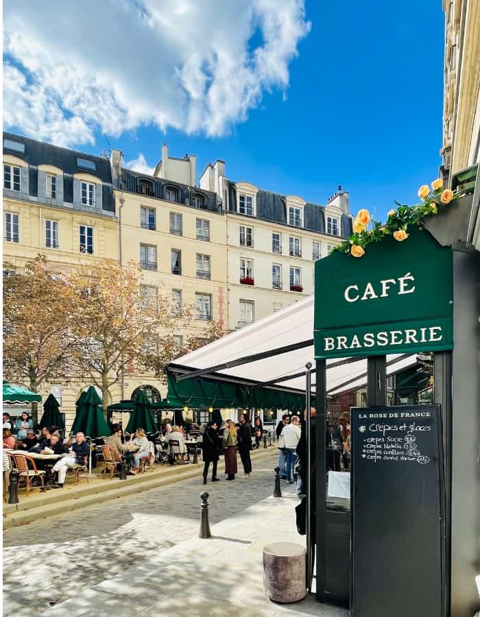 Place Dauphine on Île de la Cité in Paris. In the foreground a sign that says Café Brasserie. In the center of the square is a garden area with tables, chairs and umbrellas.