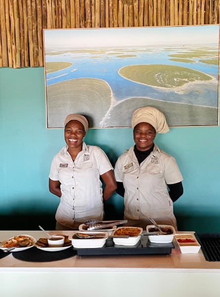Two women staff at Leroo La Tau safari lodge stand smiling in front of a lunch buffet.