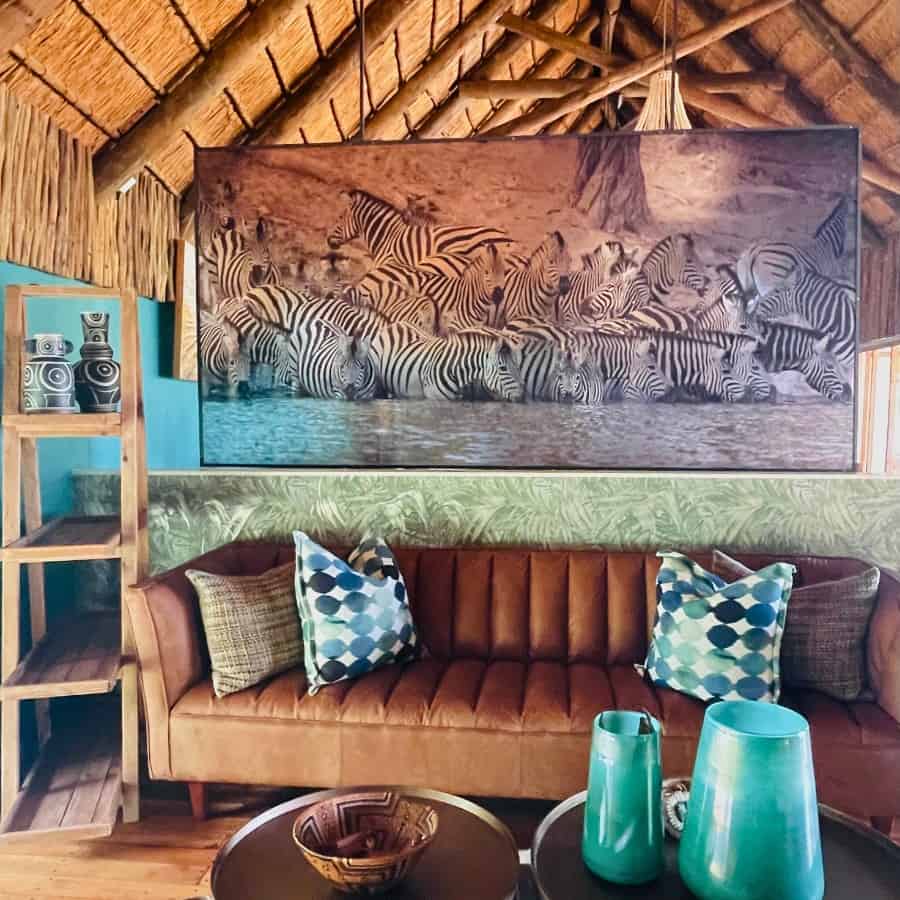 The entry area at Leroo La Tau Safari Camp in Botswana. It features a vaulted thatched roof at sitting area and a dramatic photo of the zebra migration. Colors are brown with turquoise accents.