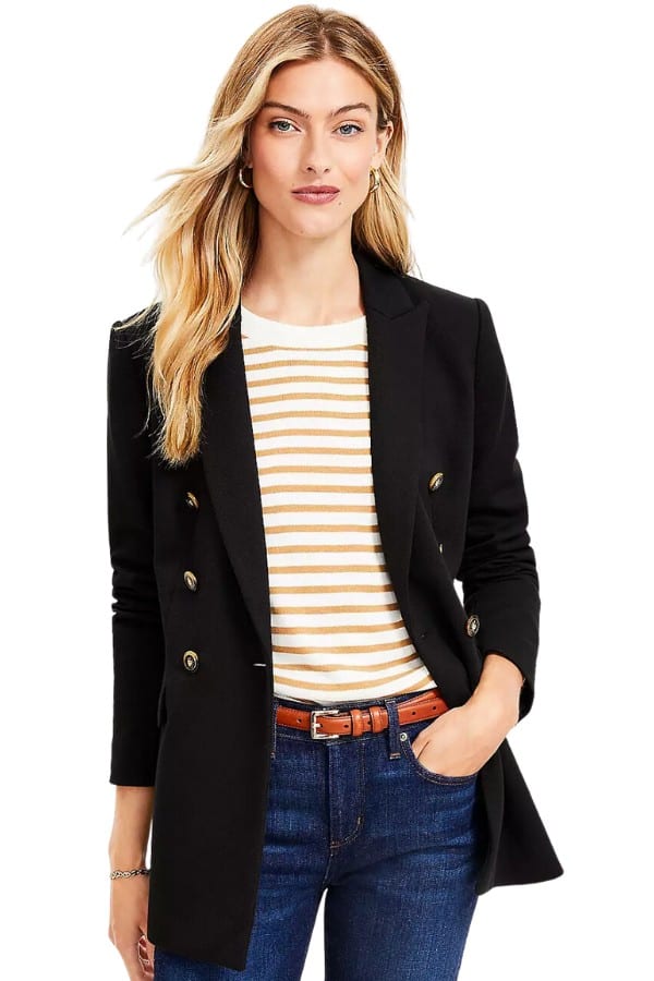 A slender woman in her early 40s is dressed in a black double-breasted blazer, layered over a yellow and white striped shirt. She is also wearing blue jeans and a slender belt.