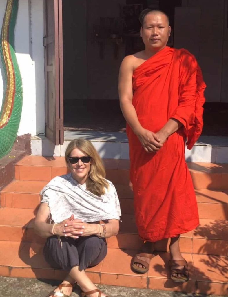 A woman sits on a stone step with a monk standing next to her. He is wearing a daffron colored robe. She is wearing pants and a tee with a scarf around her shoulders.