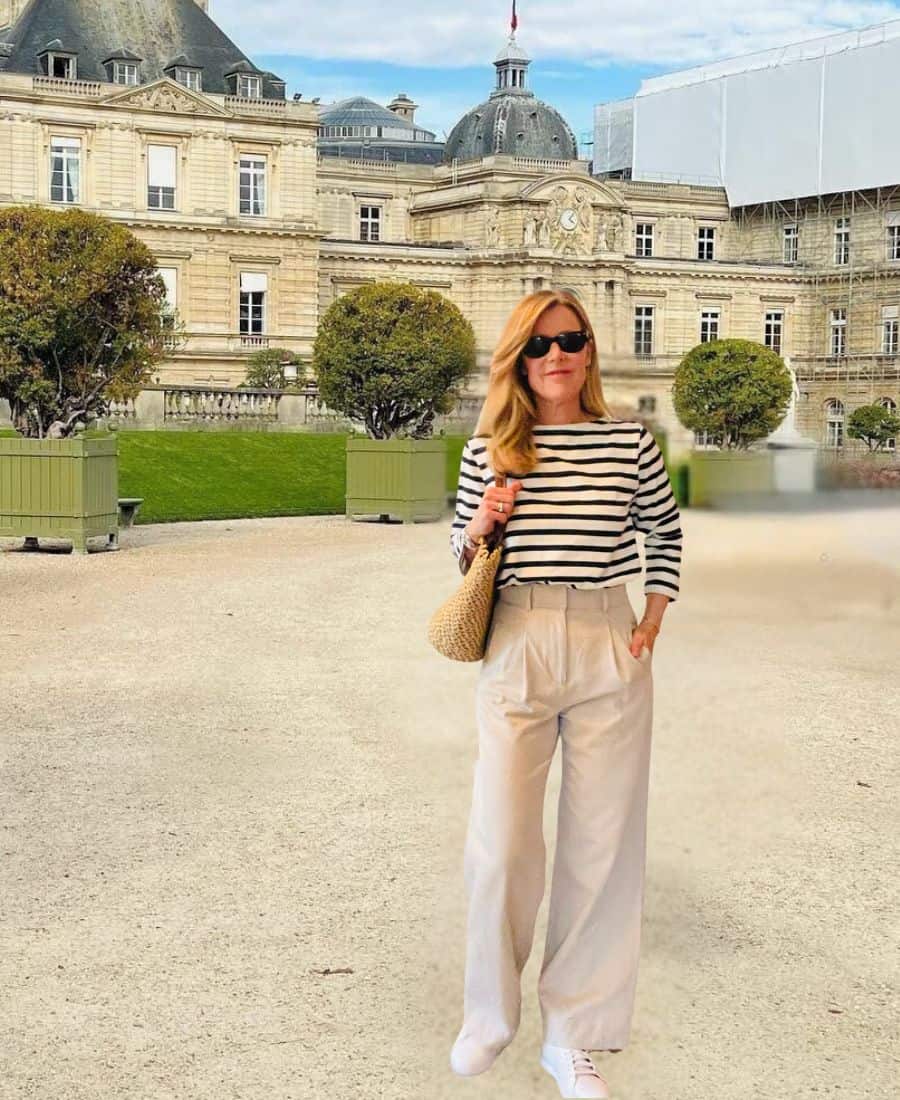 A woman stands in a Paris garden in a striped top, trousers and sneakers. She is holding a straw bag and wearing sunglasses.