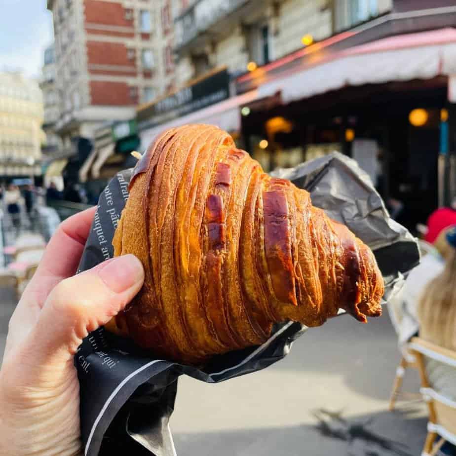A hand holds a croissant from Maison d'Isabelle bakery in Paris. A Paris street is visible beyond.