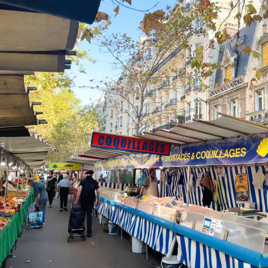 The Marché Raspail farmers market in Paris is pictured on a fall morning. Tables with blue striped decor are lined with fish and a sign coquillage (seafood in French) hangs above. A man in a black beret is pulling his cart in the distance and Haussmann architecture is visible beyond the market.