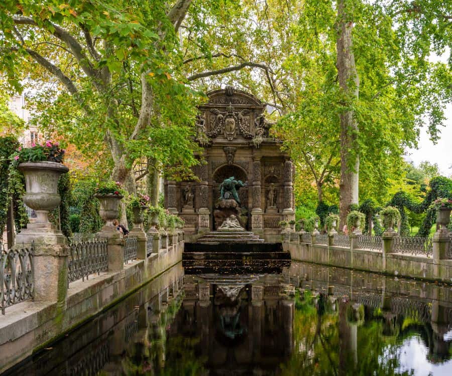 The Medici Fountain in the Luxembourg Gardens of Paris France. Pictured at the end of the fountain is the sculpture Polyphemus Surprising Acis and Galatea, by Auguste Ottin. It was added to the fountain in 1866. Urns with flowers line both sides of the fountain and trees are reflected in the water below.