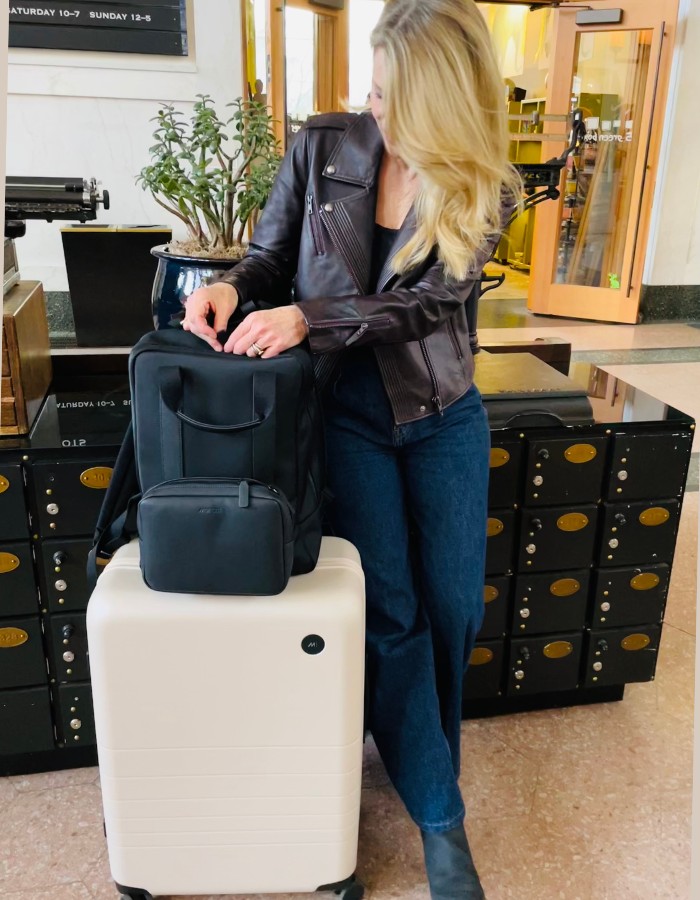 Susan Heinrich at Denver Union Station with Monos travel products: the Monos Check-In Medium roller bag in desert taupe and the Metro backpack in black. Monos luggage is featured in her guide, gifts for travel lovers.