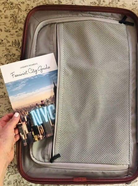 Someone slides a thin booklet called Feminist City Guide into an interior pocket of a Monos Carry-On bag