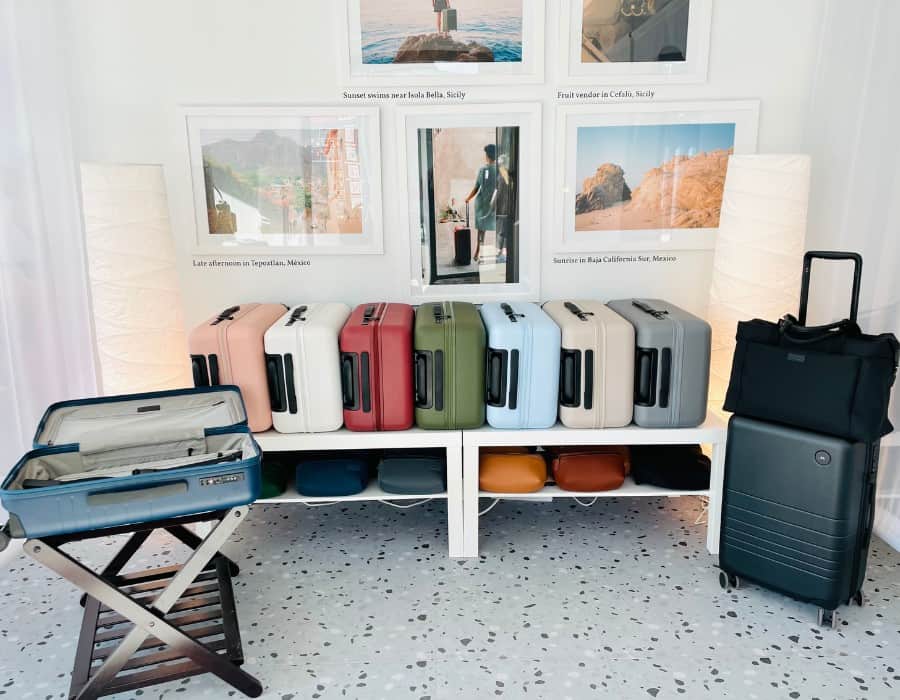 A display of Monos luggage at a popup store in Toronto's Stackd Market. Pictured are a row of Monos Carry-On bags in 7 colors as well as various other luggage items. Beyond the luggage is an artwork collage in pastel colors.