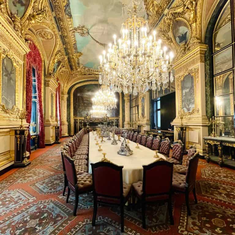 The dining room at the Napoleon Apartments at the Louvre in Paris. A Paris hidden gem recommended by Paris blogger, Susan Heinrich. A dining table that seats 40 is set in an opulent room, decorated as it was in 1865 when the Louvre was a palace.