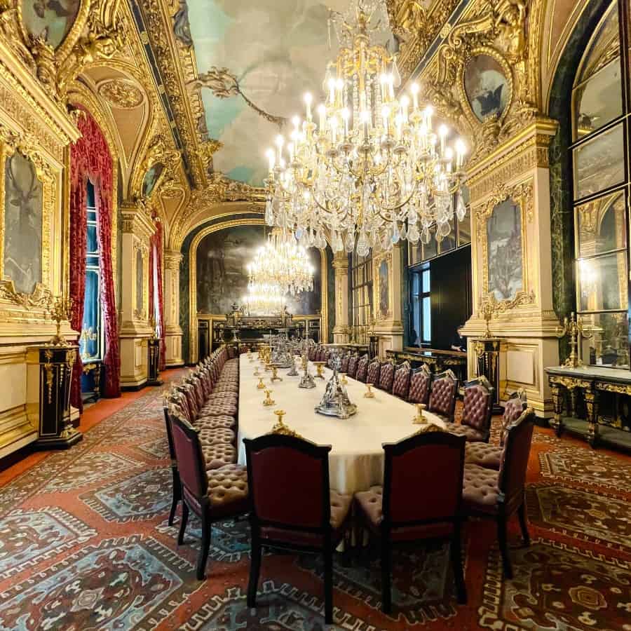 The dining room at the Napoleon Apartments at the Louvre in Paris. A Paris hidden gem recommended by Paris blogger, Susan Heinrich. A dining table that seats 40 is set in an opulent room, decorated as it was in 1865 when the Louvre was a palace.