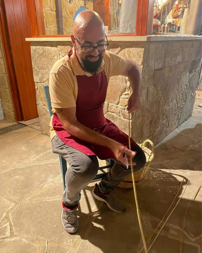 A man holds a long dry grass called a water reed. He is seated on a chair.