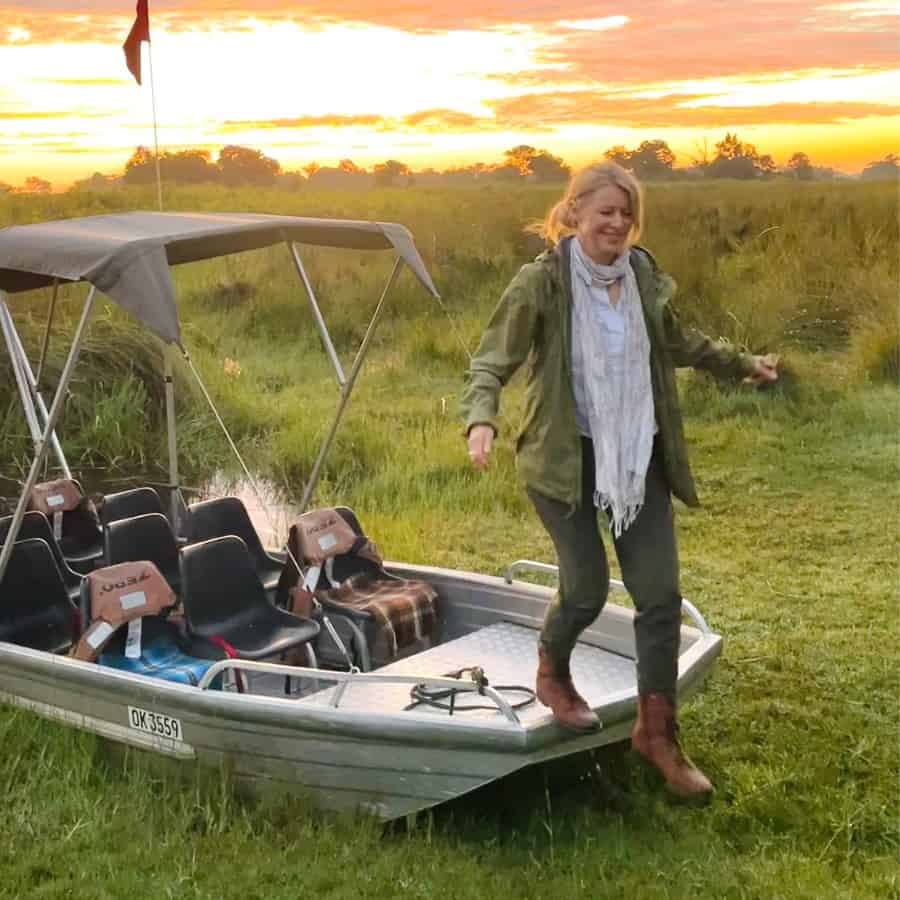 Susan Heinrich steps off of a boat in the Okavango Delta in Botswana, with the sun rising beyond her. Her safari outfit is a pair of green pants, a green rain jacket and brown safari boots.